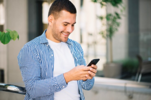 Homem jovem de camisa xadrez azul aberta com blusa branca por baixo com o telefone na mão, olha para ele sorrindo.