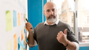 Homem adulto careca e de barba branca em frente a uma lousa com post-its coloridos. Ele segura uma caneta com uma mão e com a outra ergue o polegar.