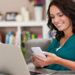 Mulher jovem de cabelos ondulados nos ombros sentada mexendo em seu celular em frente a um notebook. Ela usa uma blusa azul petróleo.